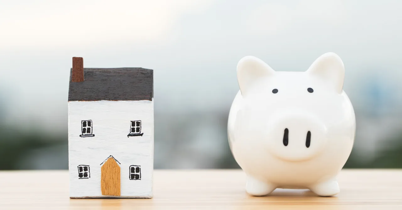 木のテーブルに並んで置かれた白い豚の貯金箱と小さな家の模型。日銀の利上げが預金と住宅ローンに与える影響を解説する記事のヘッダー画像