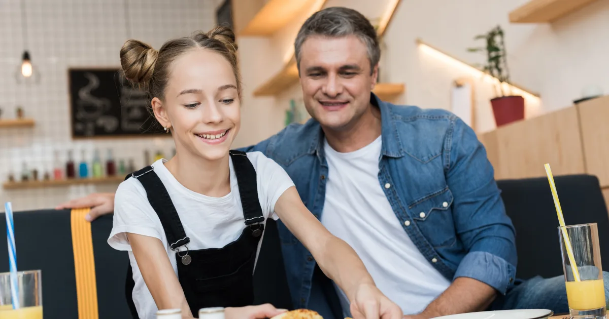 明るいカフェのテーブルで、父親と娘が笑顔で料理を取り分けている様子