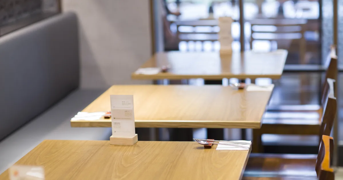 客のいない飲食店の店内に、メニュースタンドとカトラリーを置いた木製のテーブルが並んでいる様子