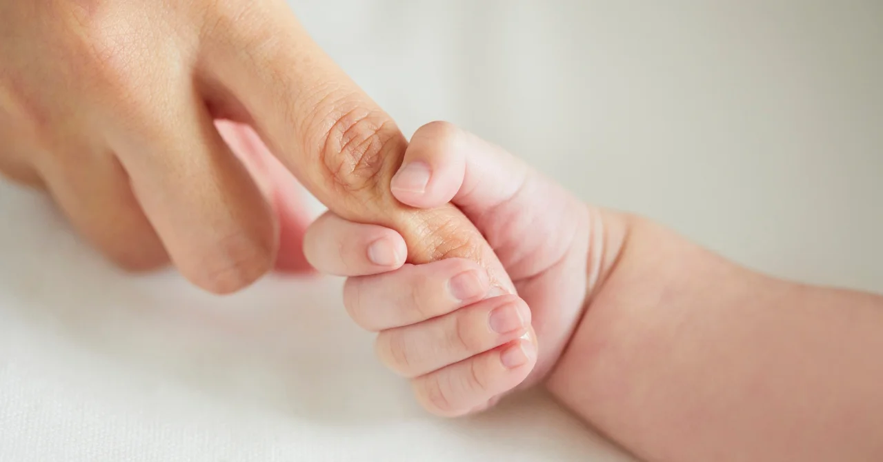 白い布の上で、赤ちゃんの小さな手が大人の指を握っている様子
